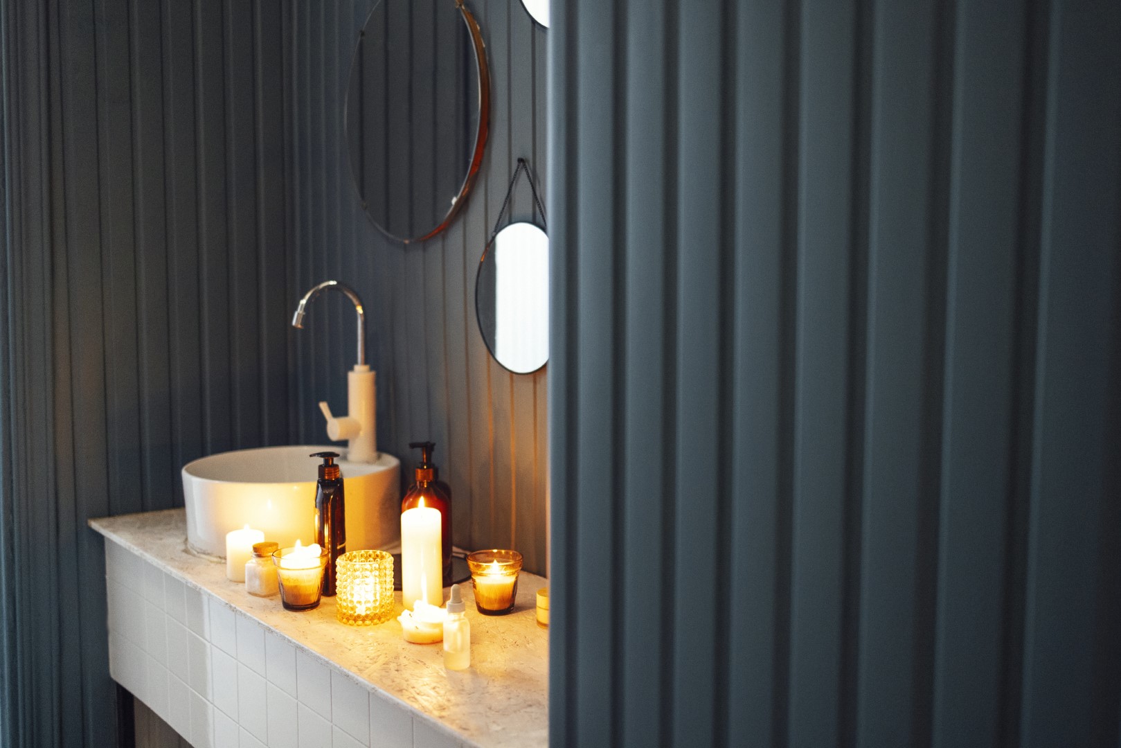 Stylish modern bathroom with mirrors, sink and candles. Dark blue walls and light marble countertops. Romantic mood with lit candles and cosmetics. Self care concept. Home spa products. Copy space.
